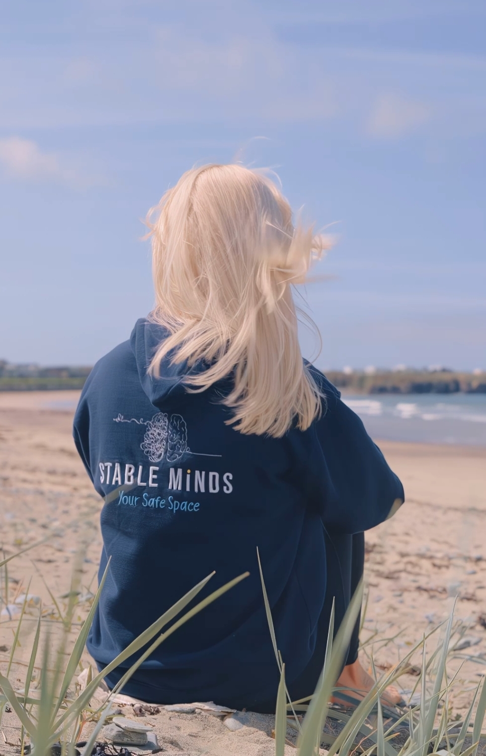 Milly sat on a beach looking out to sea, wearing a Stable Minds hoodie